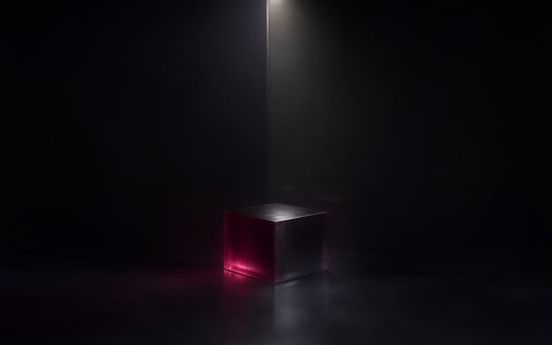 Petit cube de verre teinté de bordeaux posé dans la pénombre d'un sol sombre, à l'écart d'un fin rai de lumière blanche tombant verticalement en arrière-plan, le reste de la scène plongé dans le noir