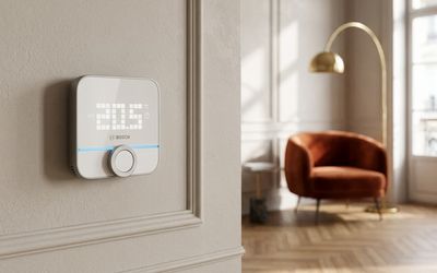 Thermostat Bosch BTH-RM blanc fixé sur un mur beige affichant 20,5°C avec sa molette rotative et sa barre LED bleue, en arrière-plan un salon avec un fauteuil en velours terracotta, un lampadaire doré et un parquet en chevrons