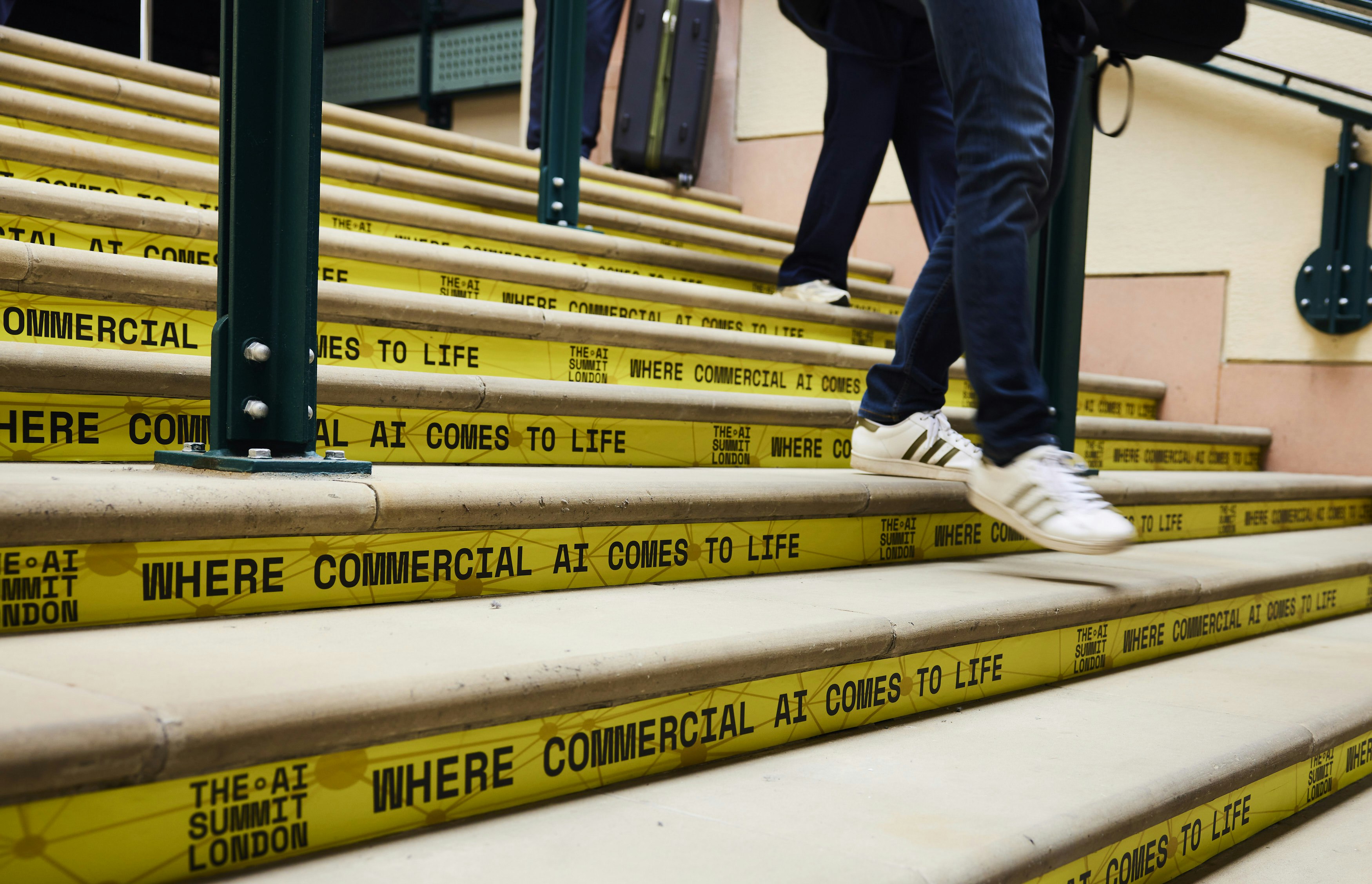 Photographie en contre-plongee d'un escalier de pierre dont chaque marche est habillee d'un adhesif jaune portant le slogan « WHERE COMMERCIAL AI COMES TO LIFE » et le logo « THE AI SUMMIT LONDON » ; au premier plan les jambes d'une personne en jean et baskets blanches qui descend les marches, a l'arriere-plan une autre personne tirant une valise