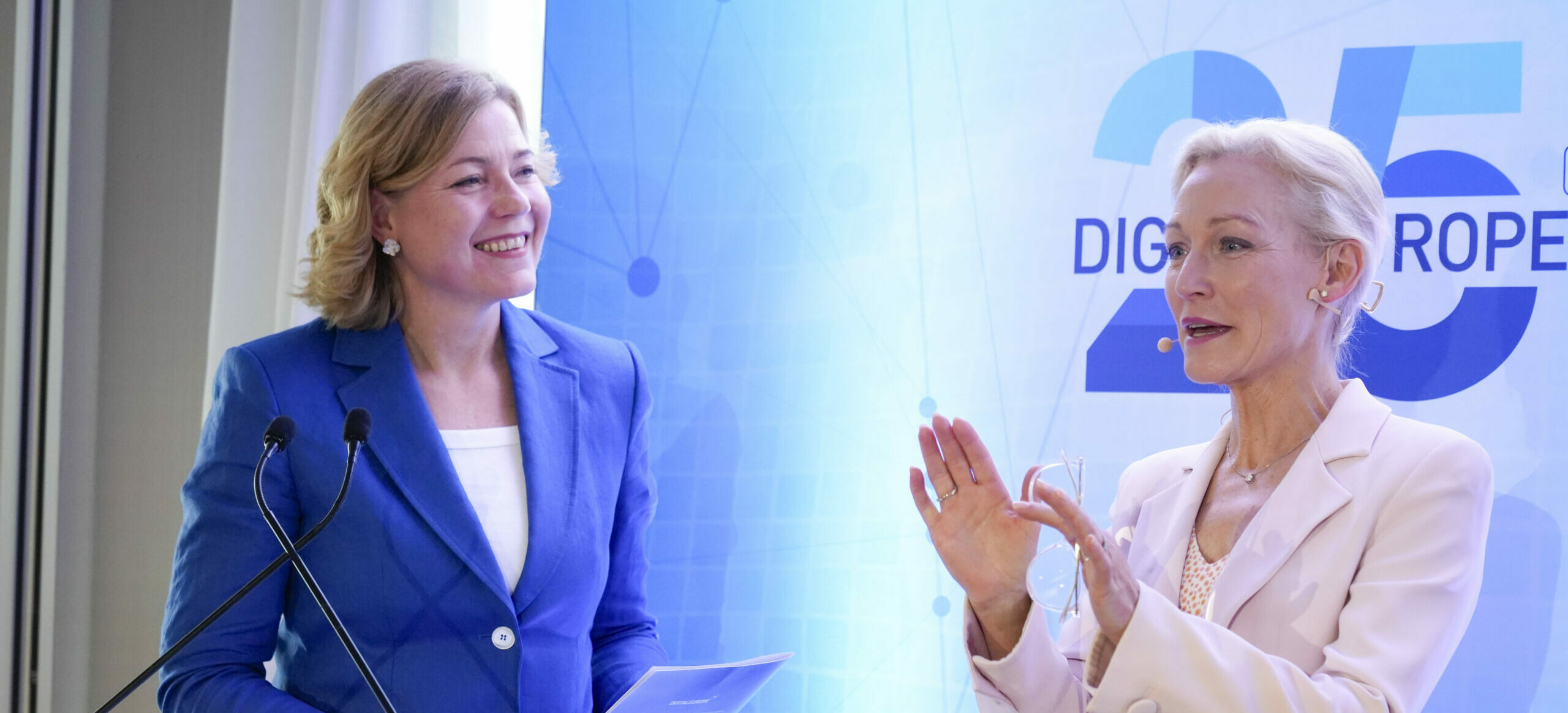 Deux femmes en tailleur sur scène, l'une en bleu et l'autre en beige, panneau DIGITALEUROPE 25 en arrière-plan bleu