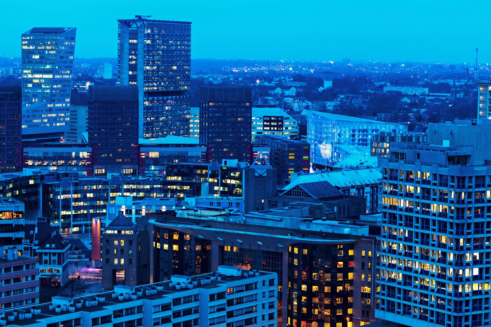 Vue aerienne du quartier d'affaires de Lille a la tombee de la nuit, dans une dominante bleue : au premier plan les toits d'immeubles residentiels, au second plan des tours de bureaux aux fenetres eclairees, a l'arriere-plan la ville qui s'etend jusqu'a l'horizon sous un ciel bleu profond