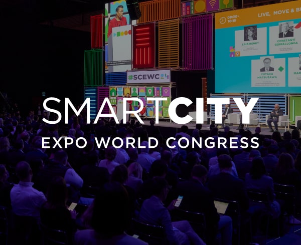 Salle de conference du Smart City Expo World Congress vue depuis le fond : au premier plan un public nombreux assis de dos dans une lumiere bleutee, au second plan une scene avec deux intervenants assis en fauteuil, a l'arriere-plan un mur de conteneurs colores et un grand ecran affichant les portraits des intervenants et le hashtag #SCEWC ; en superposition, le logo blanc « SMARTCITY EXPO WORLD CONGRESS »