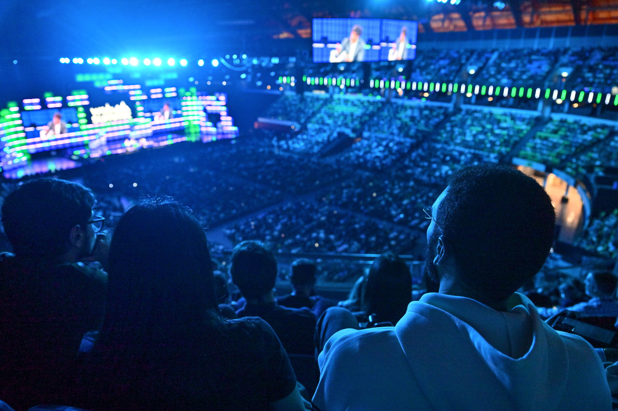 Photographie prise depuis les gradins de la MEO Arena pendant une conference, dans une lumiere bleue : au premier plan plusieurs spectateurs vus de dos et de profil, au second plan la fosse remplie de public et la scene centrale eclairee de bandeaux verts et bleus, a l'arriere-plan les tribunes bondees et un ecran geant diffusant l'image de deux intervenants