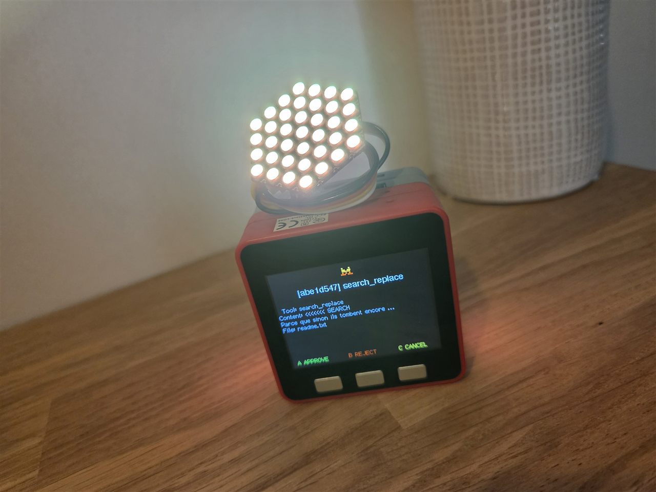 Photo du boîtier rouge M5Stack Fire posé sur une table en bois clair, écran affichant une demande d'approbation « [abe1d547] search_replace » sur le fichier readme.txt avec les libellés A APPROVE en vert, B REJECT en orange et C CANCEL en vert, une matrice LED hexagonale brillant en blanc au-dessus du boîtier, vase tressé blanc en arrière-plan