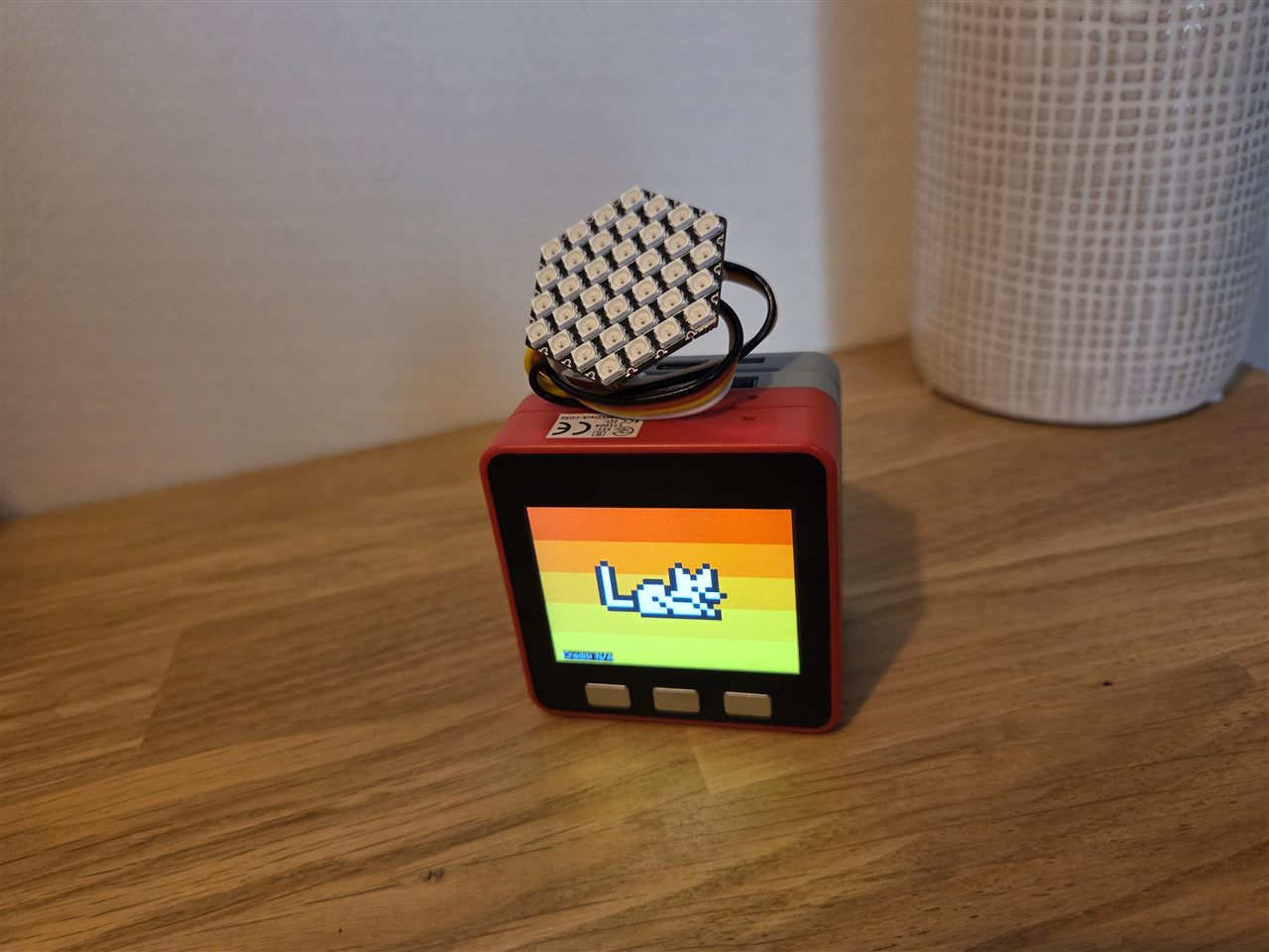 Photo du boîtier rouge M5Stack Fire posé sur une table en bois clair, écran affichant le Chaton Fat blanc en pixel art couché sur un fond arc-en-ciel rayé orange et jaune, une matrice LED hexagonale éteinte posée sur le dessus du boîtier avec son câble, vase tressé blanc en arrière-plan